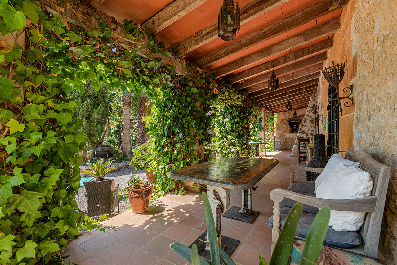 The shaded loggia: a terracotta-tiled floor, ivy along the beams, an old iron table and a bench under hanging lanterns, the garden glowing green beyond